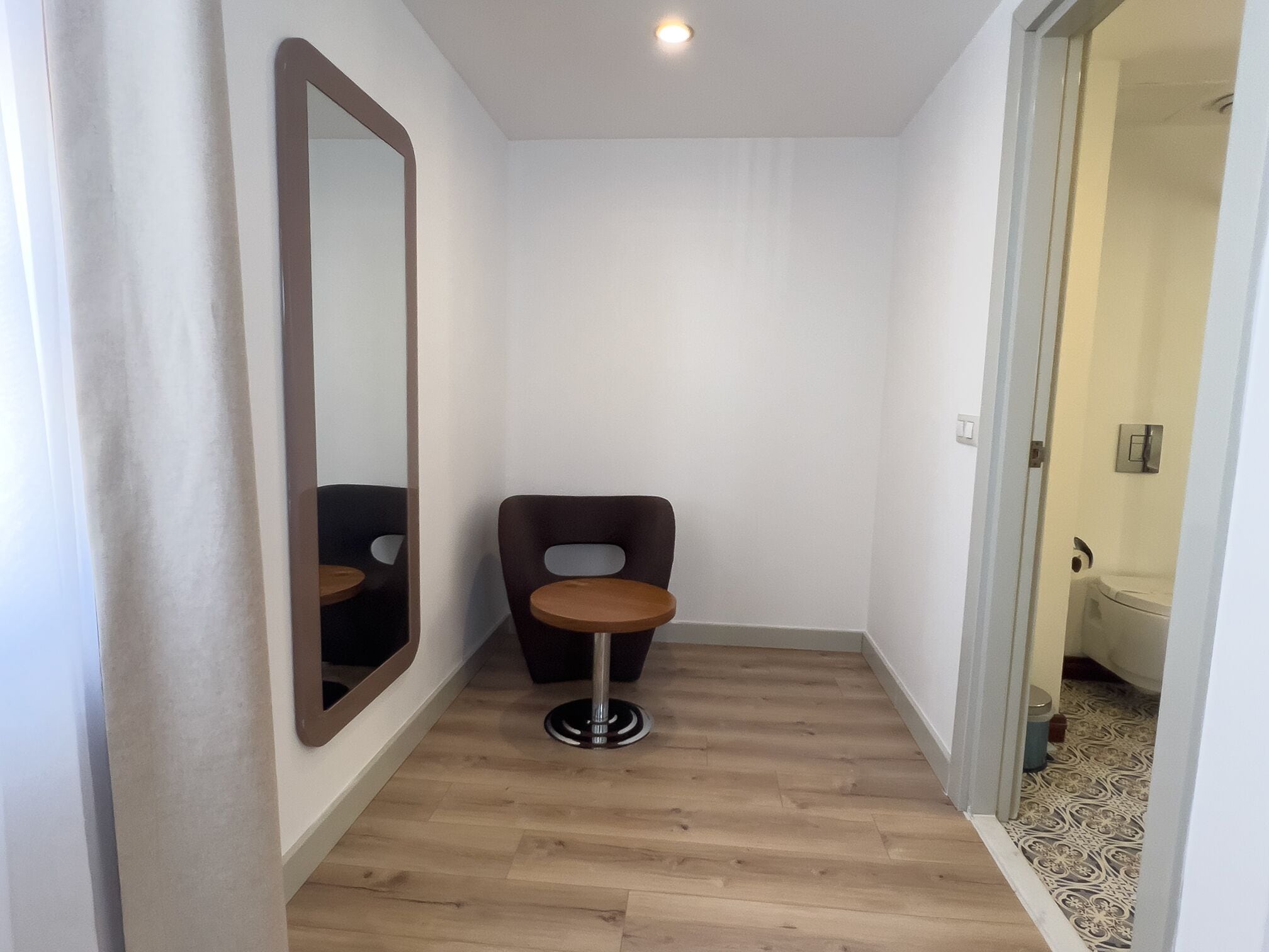 Un pasillo estrecho con suelo de madera, una silla marrón oscuro y una mesa redonda, y un espejo grande en la pared izquierda.