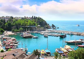 Un puerto costero con barcos amarrados, un litoral rocoso y edificios en una colina al fondo.