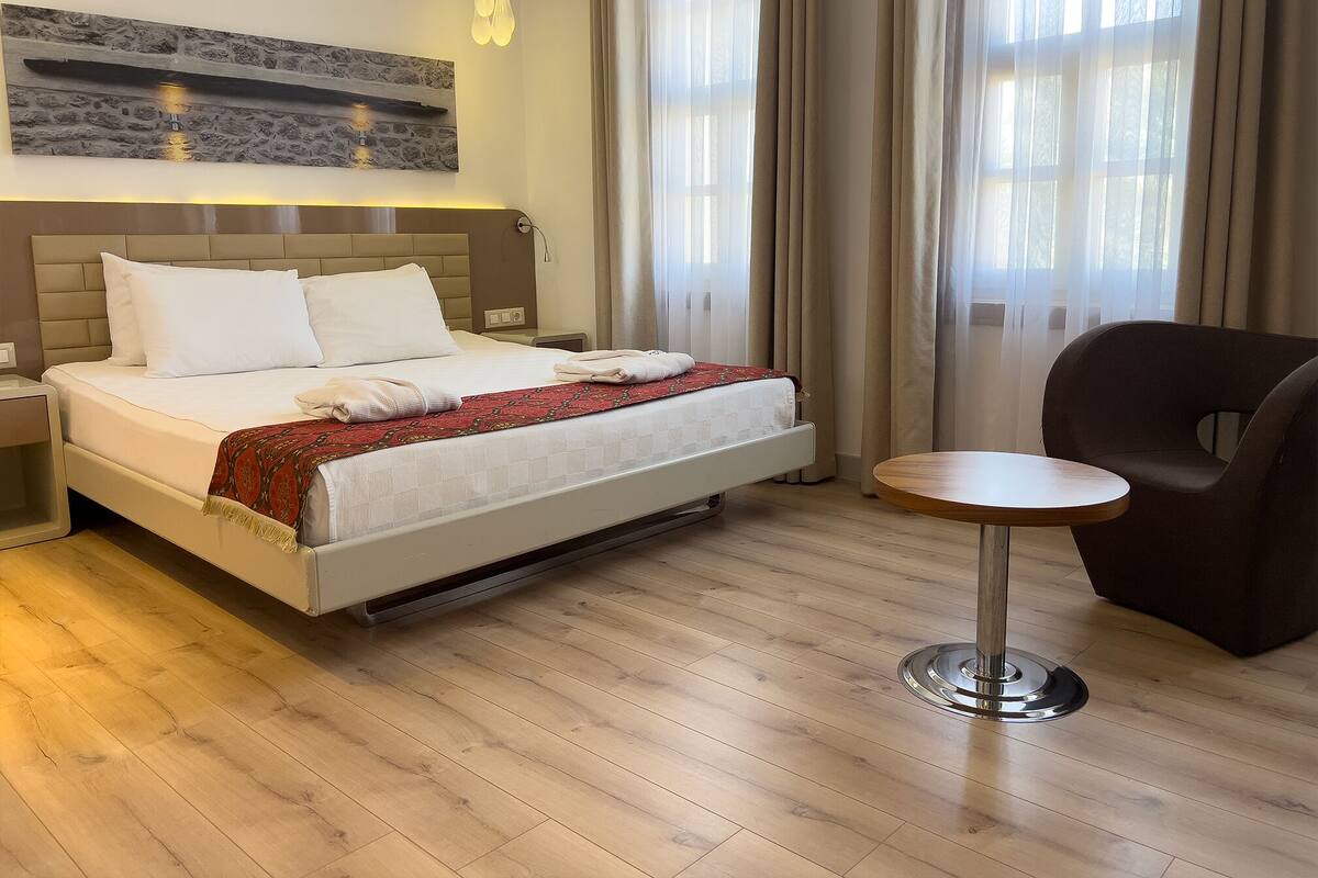 A neatly made bed with white linens and a red patterned throw, a modern chair, and a small round table in a well-lit room with wooden flooring and large windows.