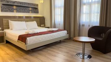 Una cama con ropa de cama blanca y un plaid rojo estampado, una silla moderna y una mesita redonda en una habitación luminosa con suelo de madera y ventanas grandes.