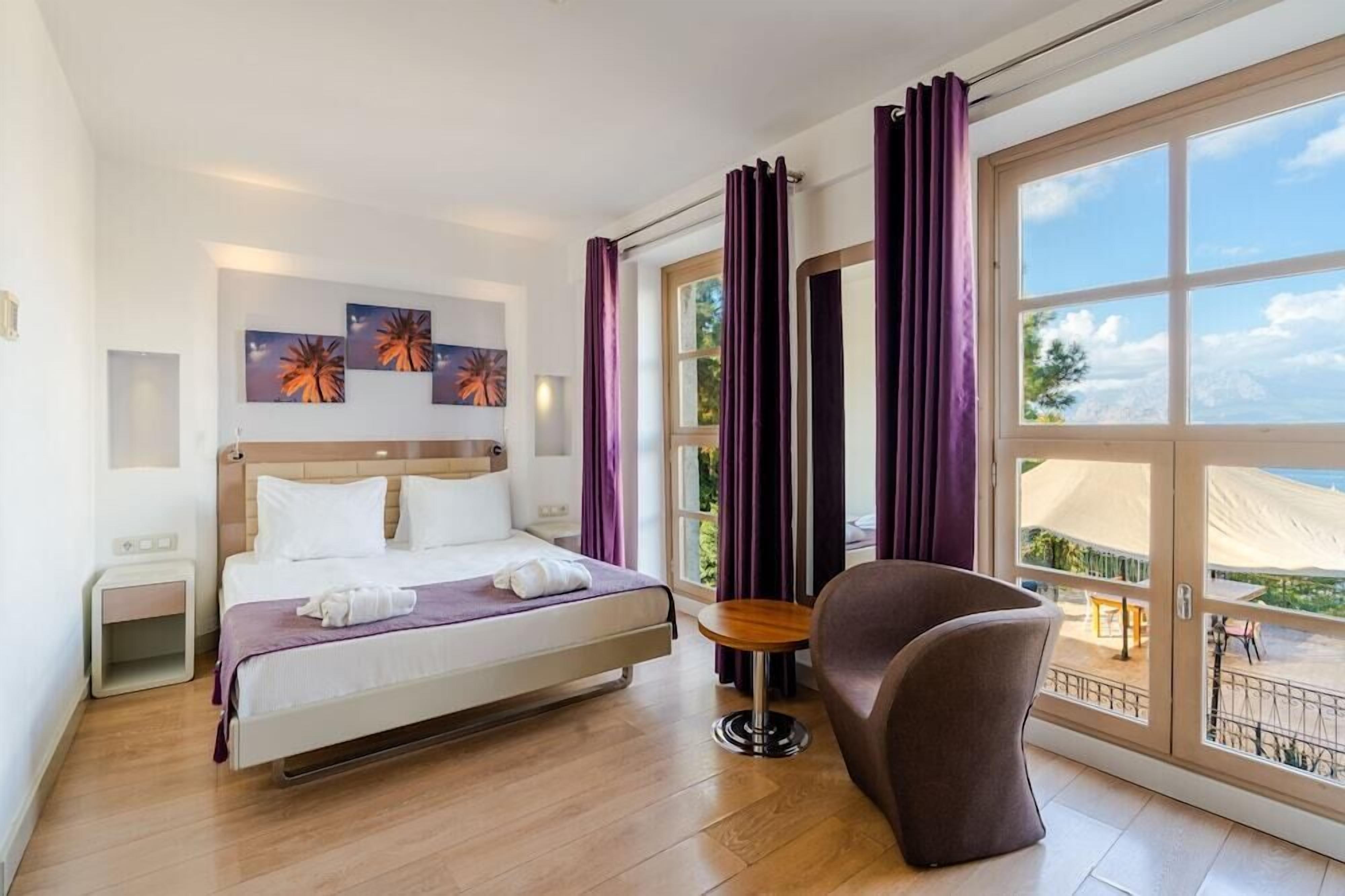 A hotel room with a bed, a small table, a chair, and a large window with a view.
