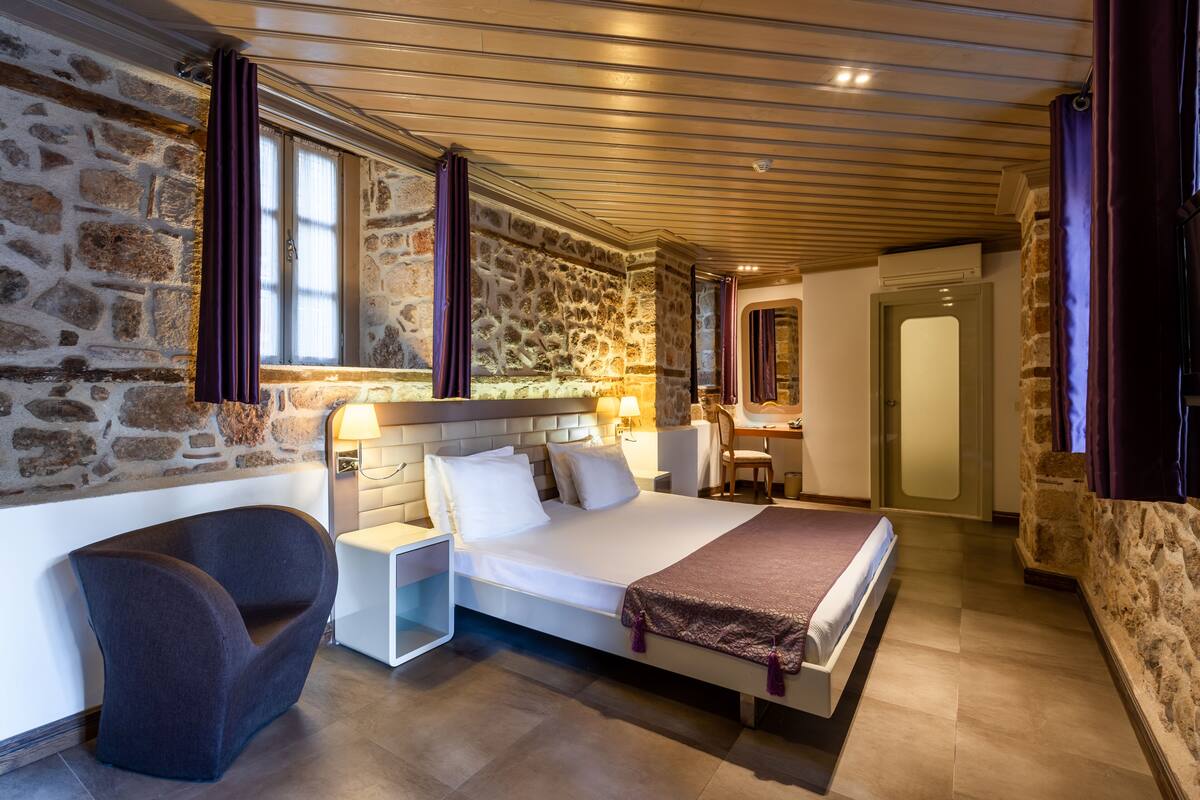 A modern hotel room with a stone wall, a bed with white linens, a curved armchair, and a desk with a lamp.