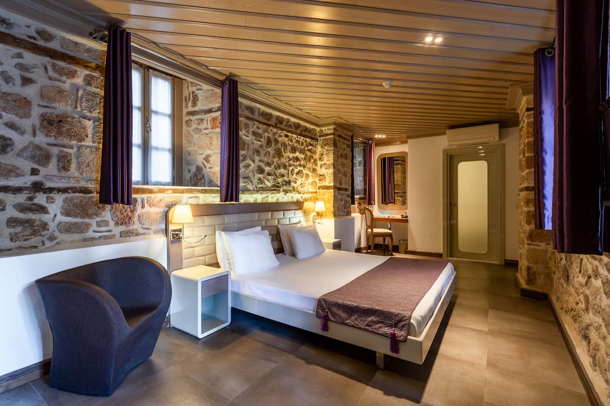 A modern hotel room with a stone wall, a bed with white linens, a curved armchair, and a desk with a lamp.