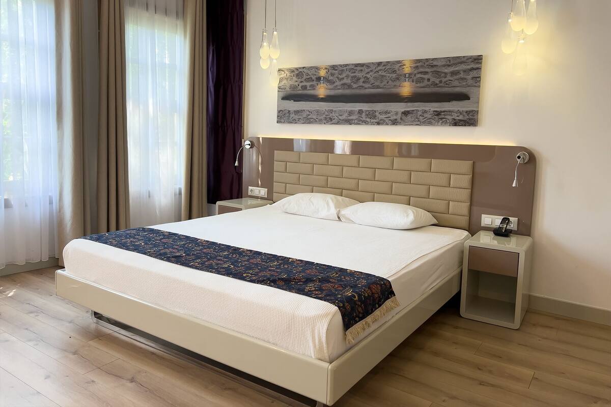 A hotel room with a large bed, a bedside table, a nightstand, a wall-mounted lamp, and a painting above the bed.