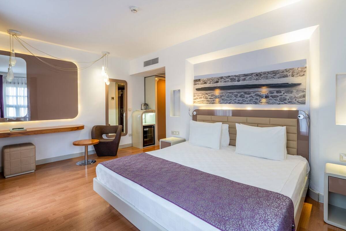 A modern hotel room with a large bed, a seating area with a chair, a small table, and a built-in bench.