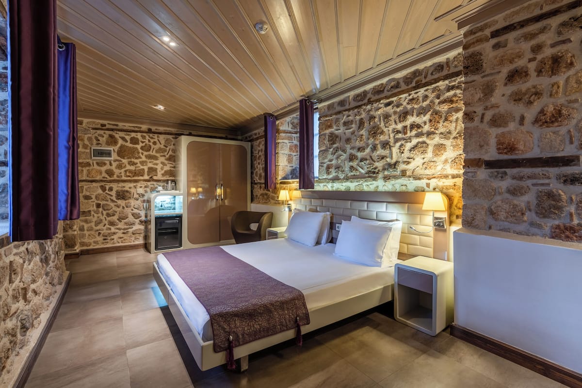 Une chambre avec un mur en pierre, un lit avec du linge de lit blanc, une table de chevet, une lampe et une petite table.