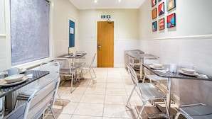A clean, well-lit kitchen with stainless steel appliances, multiple dining tables, and chairs. A wooden door with a fire exit sign above it is visible in the background. - The Park Hotel (London)