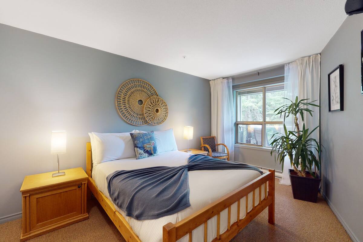 Un dormitorio con cama, mesitas de noche, una ventana con cortinas, una silla y una planta en maceta.