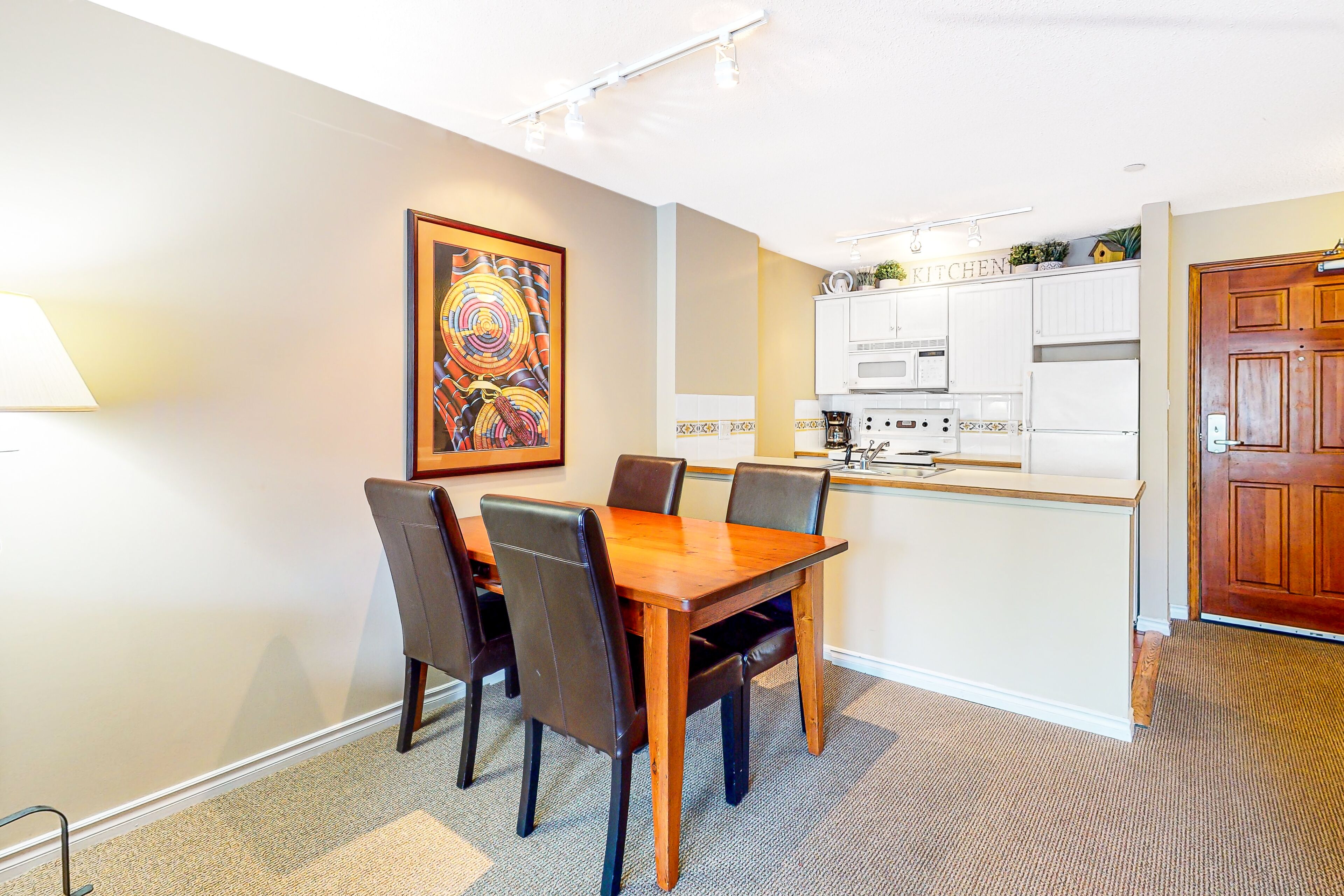 A dining area with a wooden table and chairs, a kitchen with white cabinets, and a door to the right.