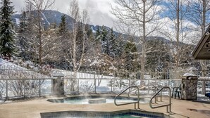 Un jacuzzi con pared de piedra y barandillas metálicas, rodeado por un paisaje nevado y árboles.