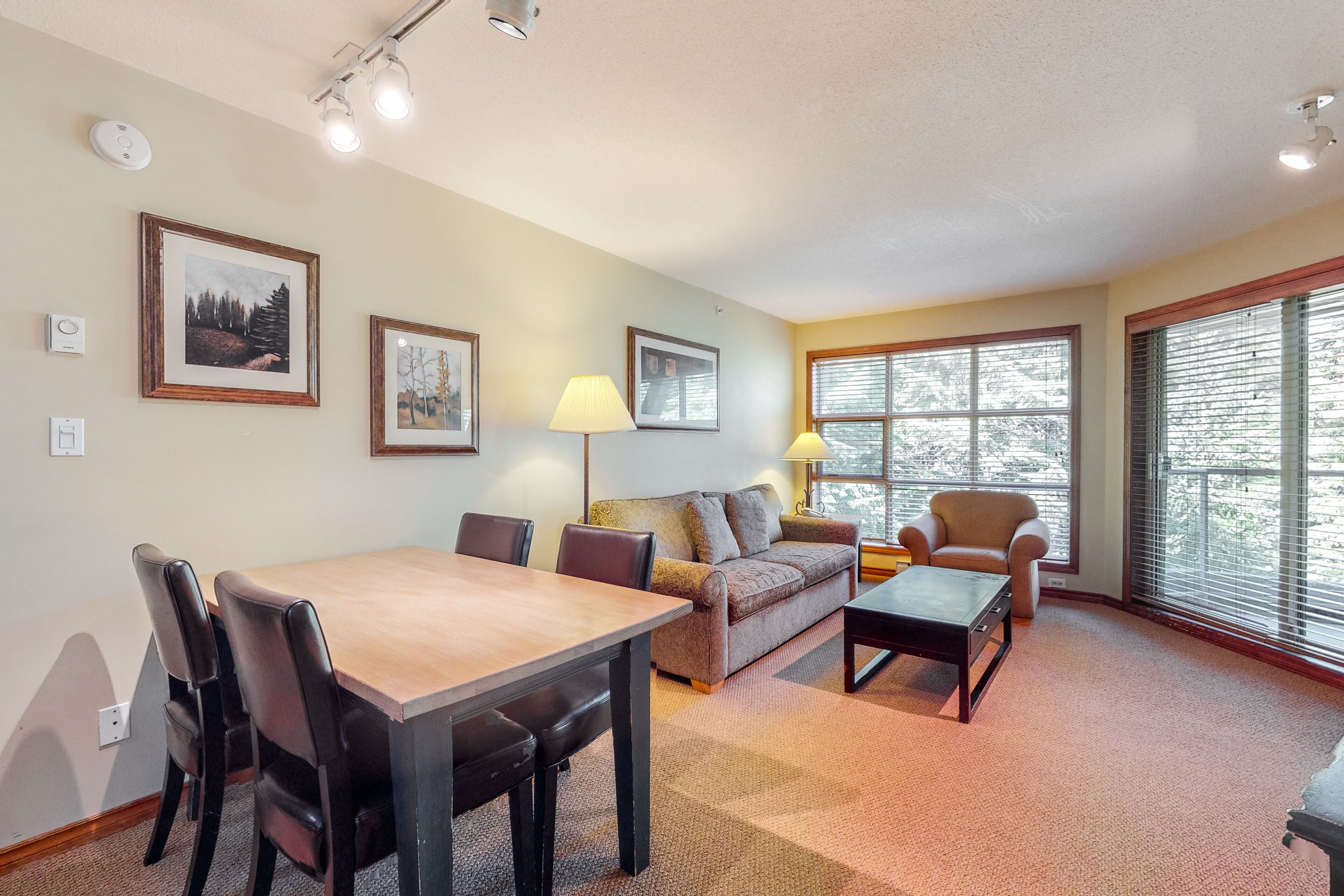 A living room with a dining table, chairs, a sofa, a coffee table, and a lamp. There are framed pictures on the wall and a window with blinds.