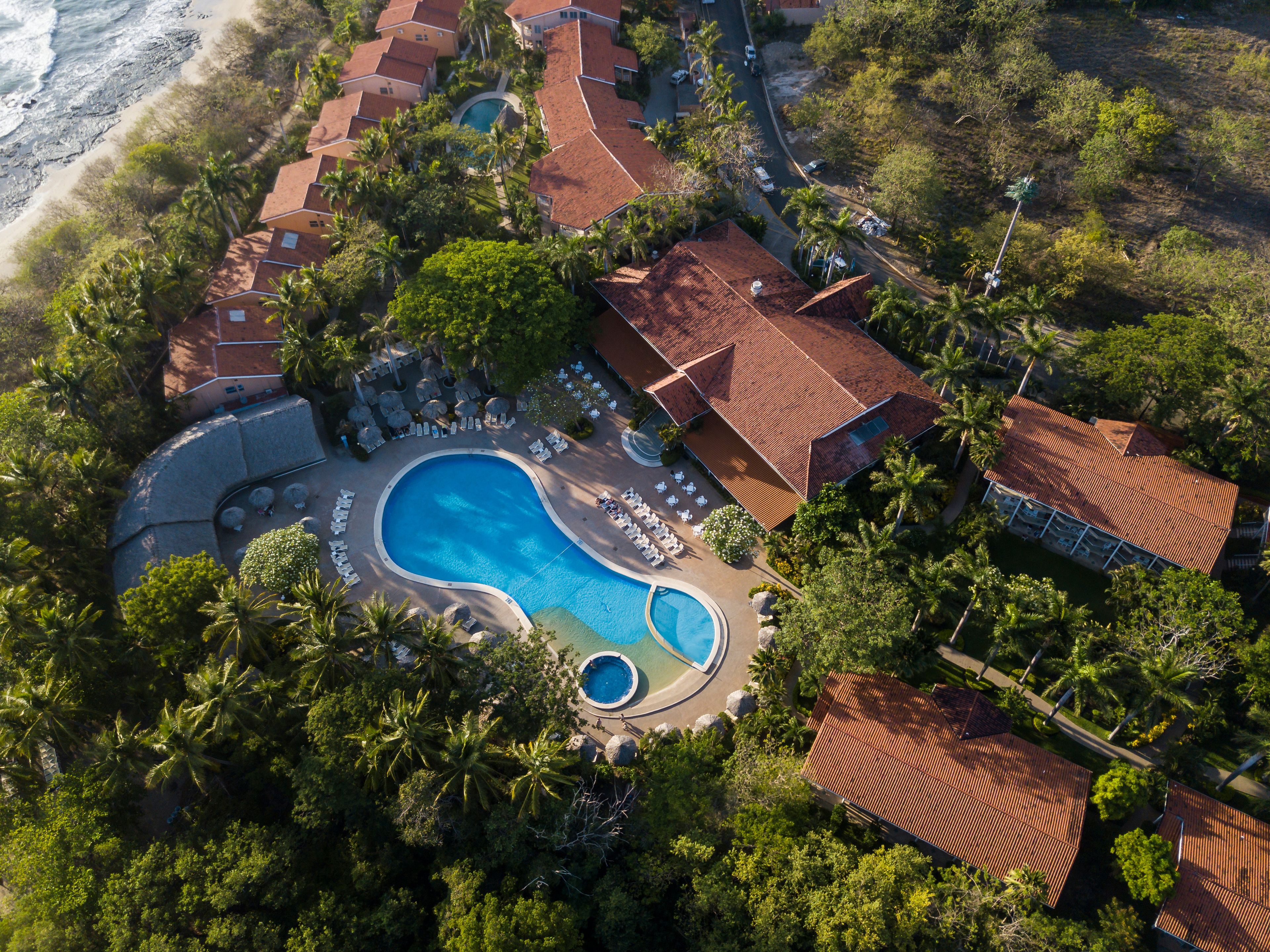 Vista aérea de un complejo turístico con varias edificaciones, una piscina grande y una playa.