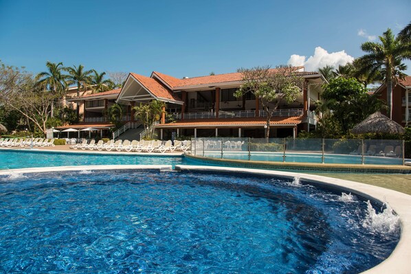 A swimming pool with lounge chairs, a building with a red roof, and palm trees. - Occidental Tamarindo - All Inclusive (Tamarindo)