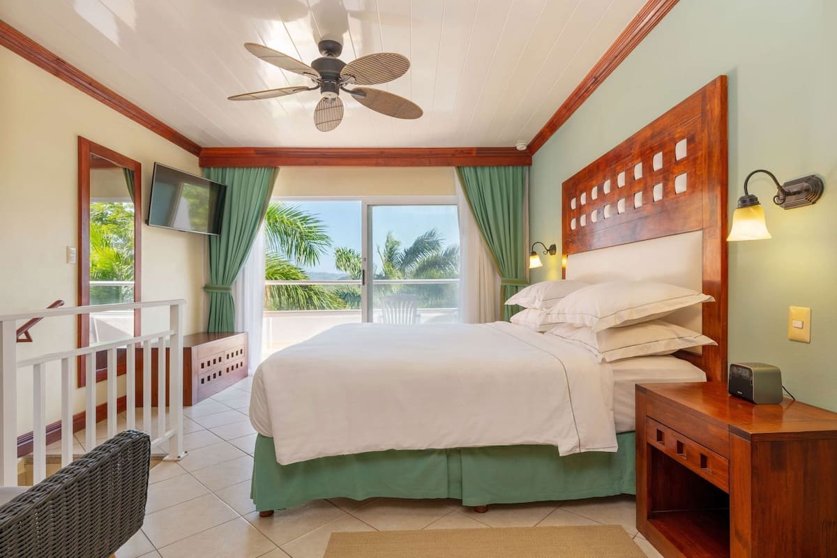 Habitación de hotel con cama, ventilador de techo, balcón con vistas, televisión y mesita de noche.