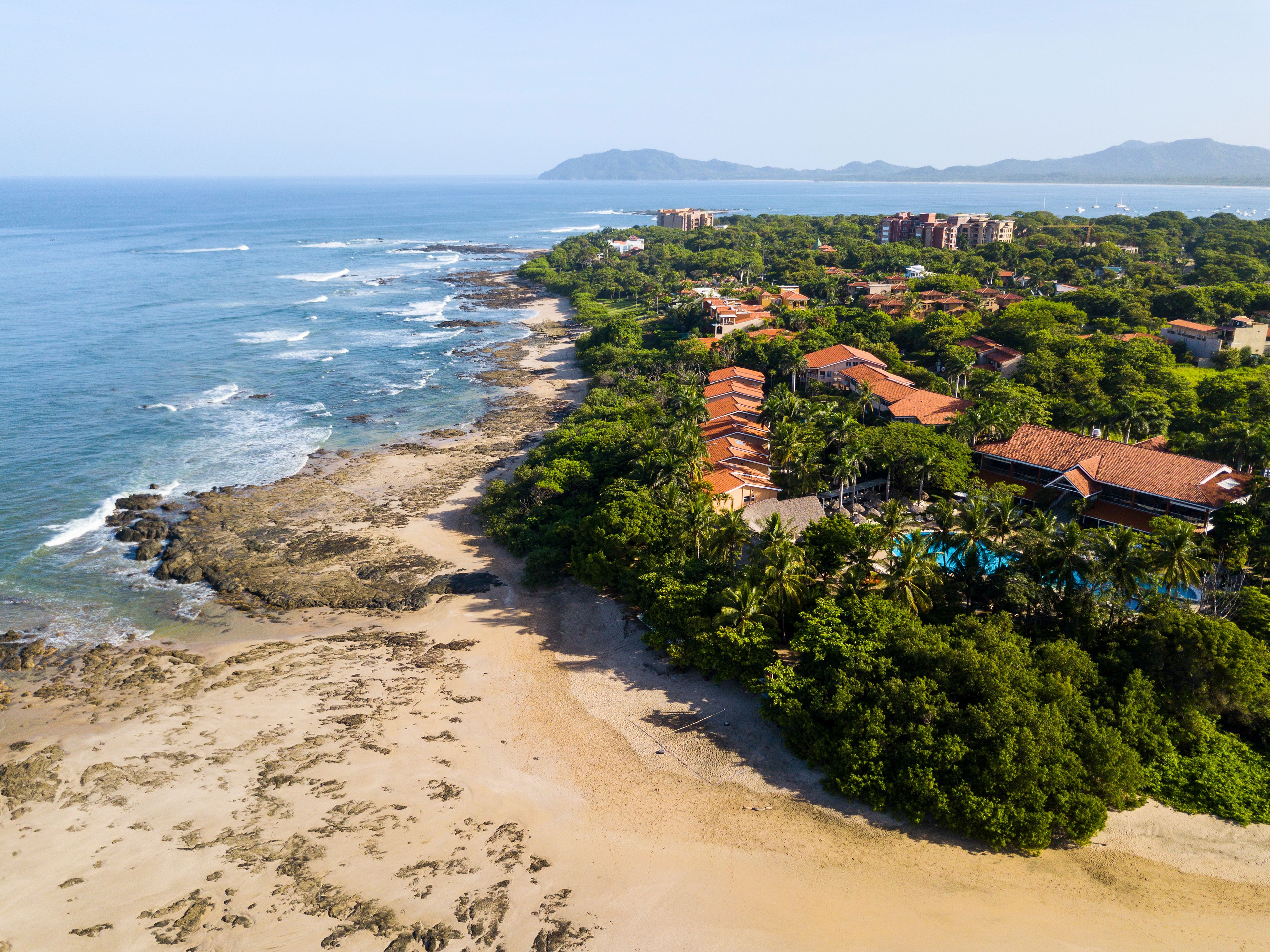 An aerial view of a coastal area with a beach, buildings, and a mountainous backdrop.