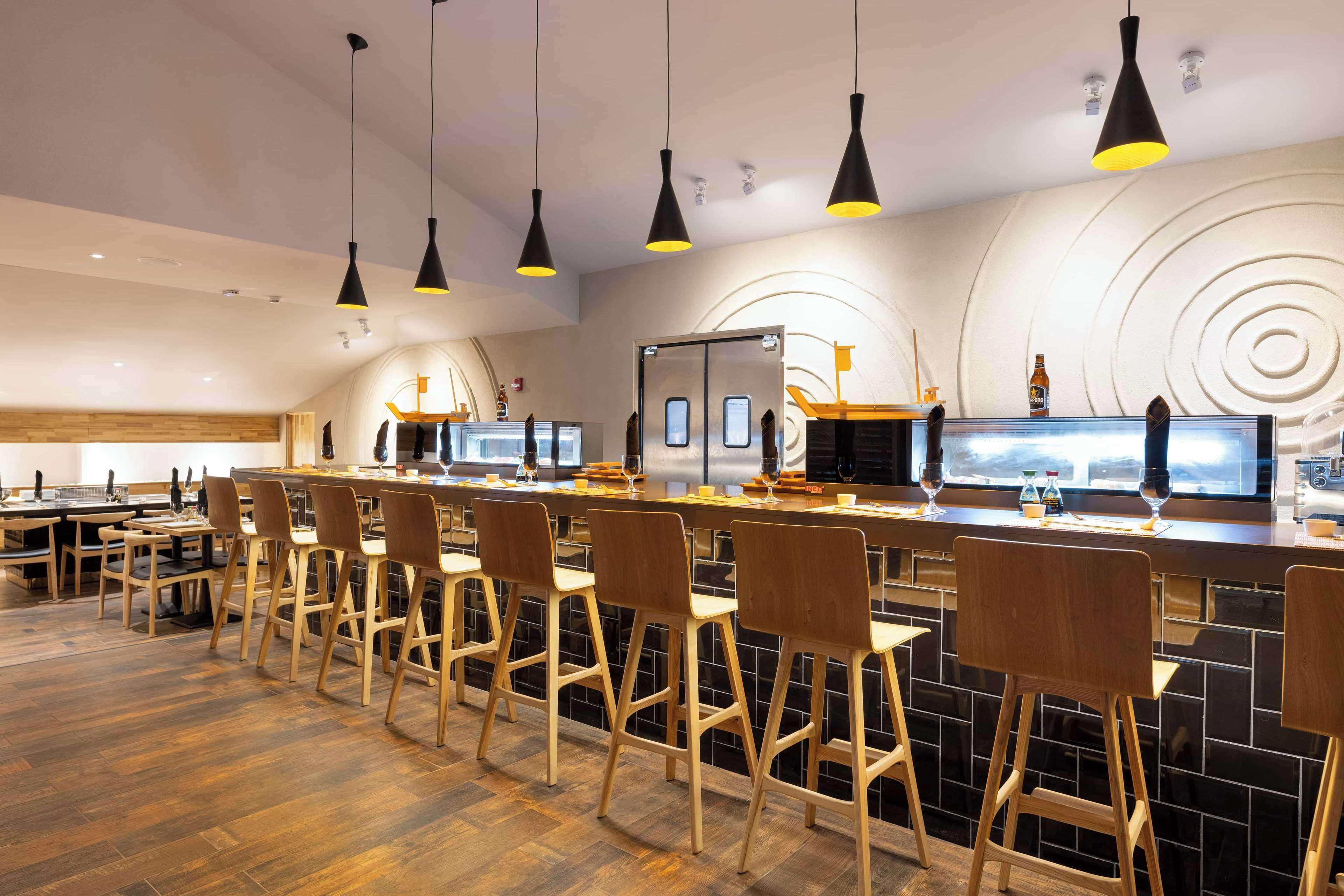 A modern bar area with wooden bar stools, black pendant lighting, and a large screen displaying a cityscape.