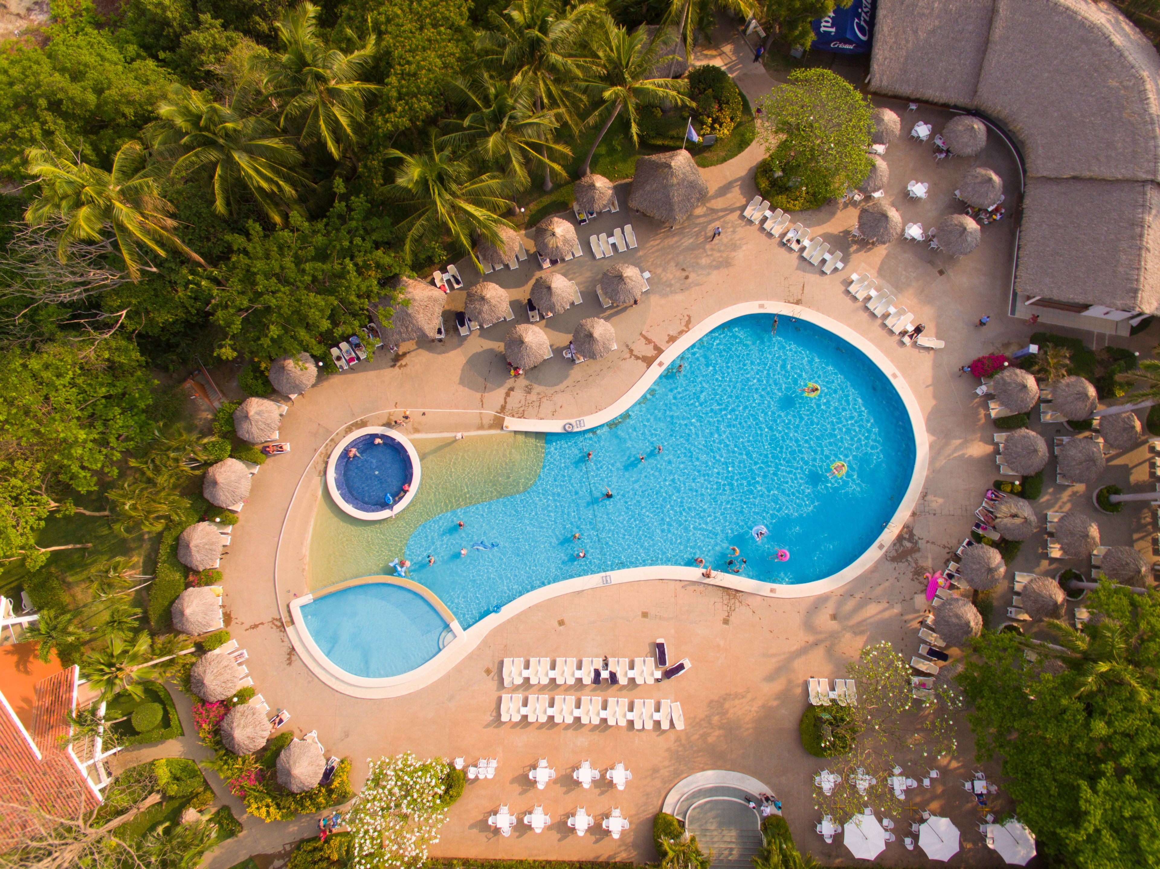 Vista aérea de um resort com uma piscina grande, espreguiçadeiras e palmeiras.