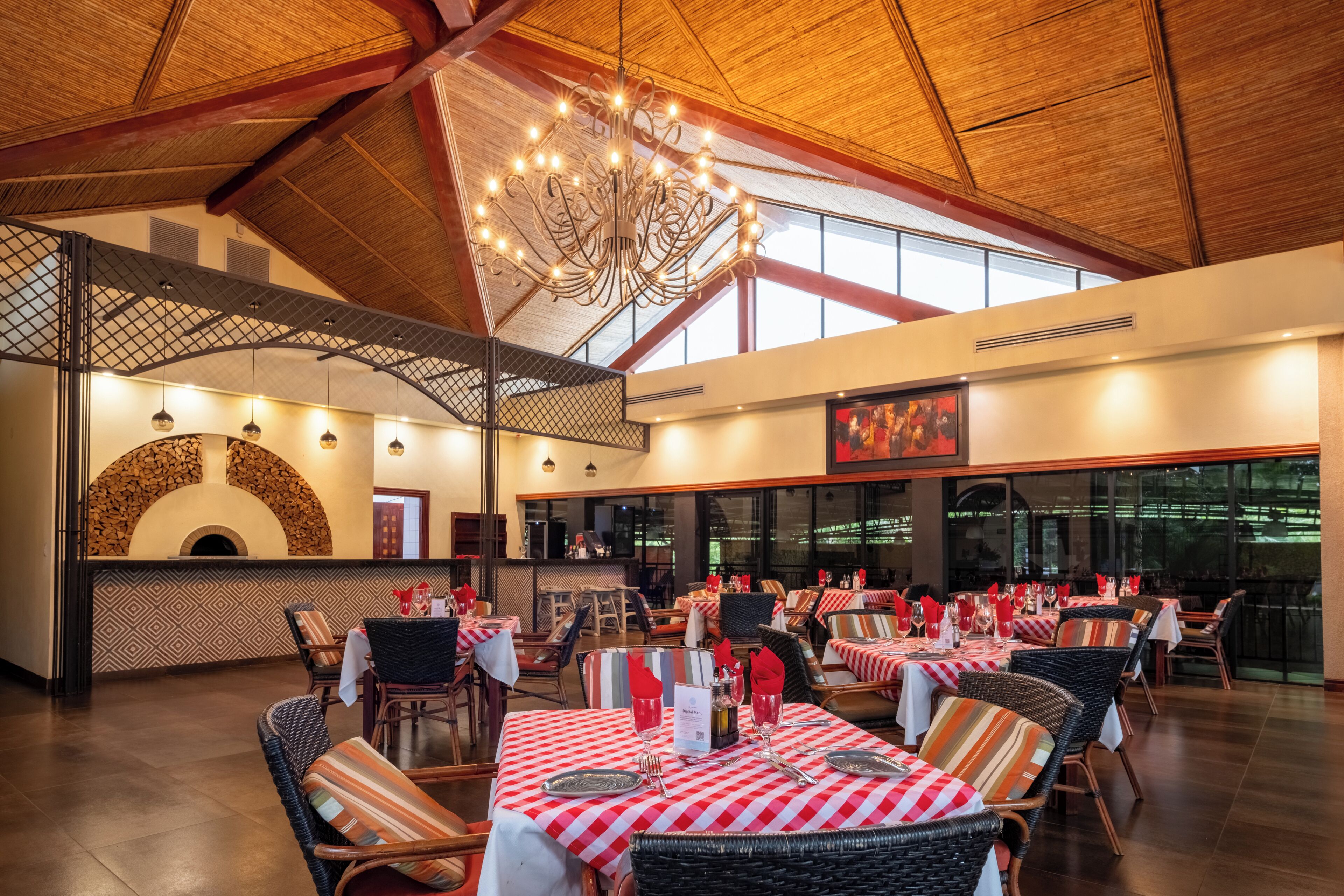Un restaurant spacieux avec de hauts plafonds, une cheminée centrale et un lustre moderne. Les tables sont dressées avec des nappes à carreaux rouges et blancs et des chaises en osier.