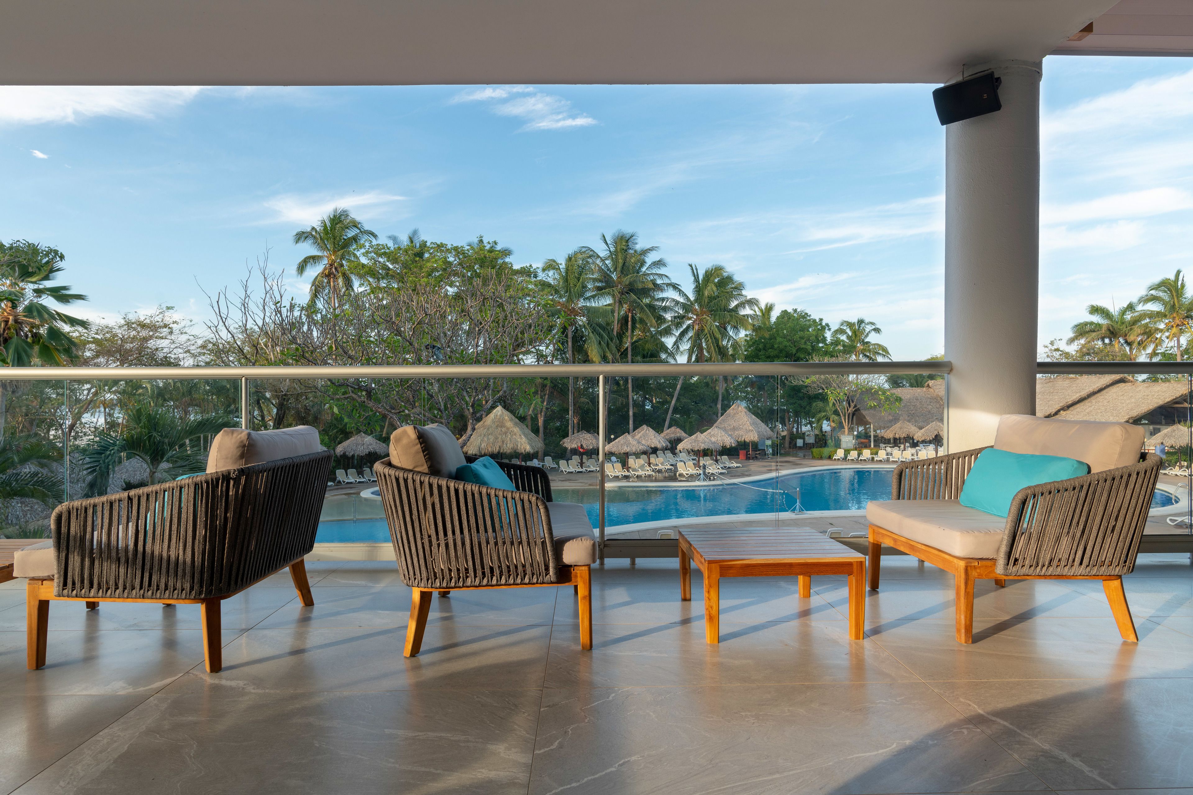 Un balcón con mobiliario de mimbre, piscina y vista a palmeras.