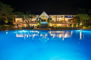 A nighttime view of a hotel with a swimming pool, outdoor seating, and a well-lit entrance.