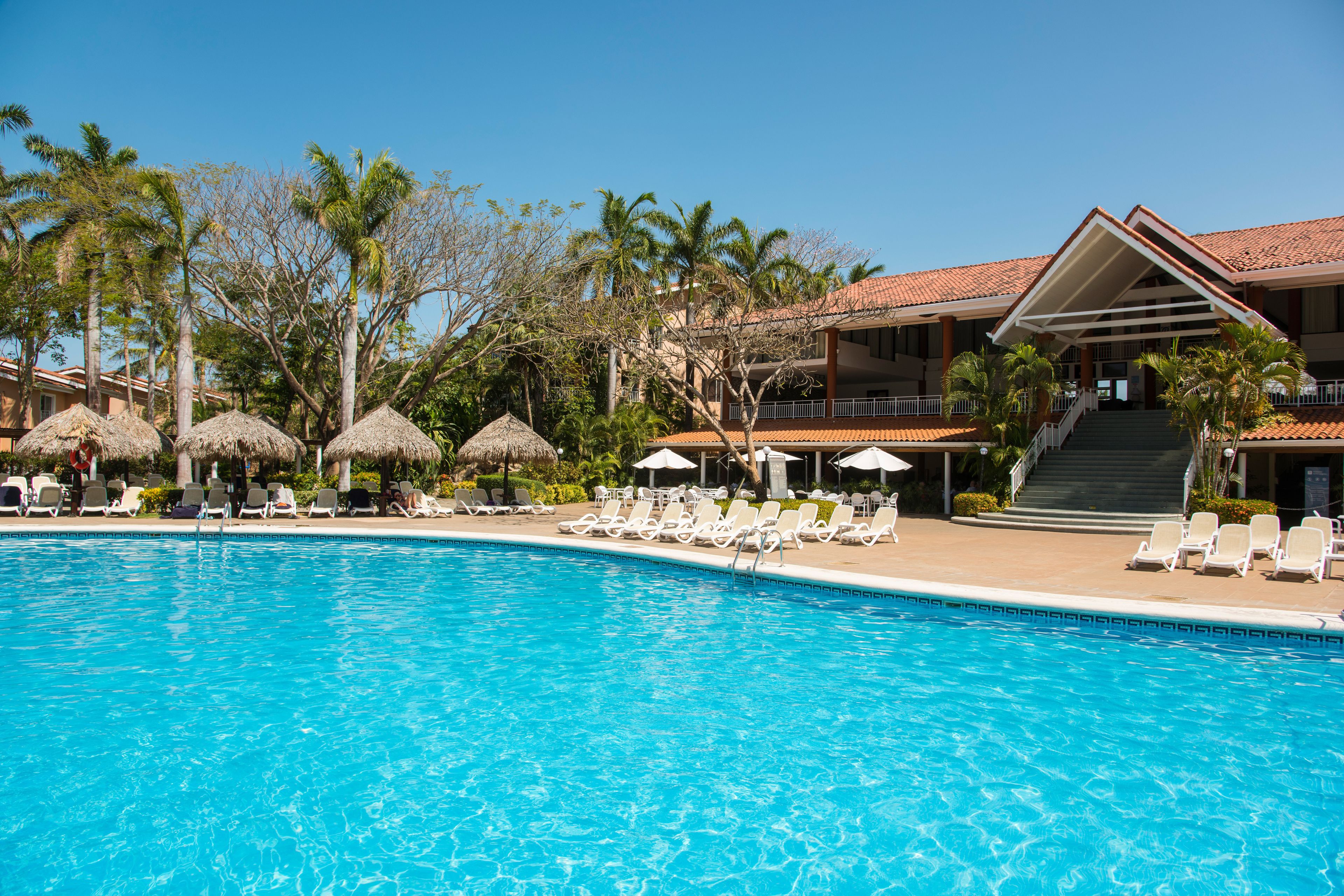 A swimming pool with lounge chairs and umbrellas, surrounded by palm trees and a building with a red-tiled roof.
