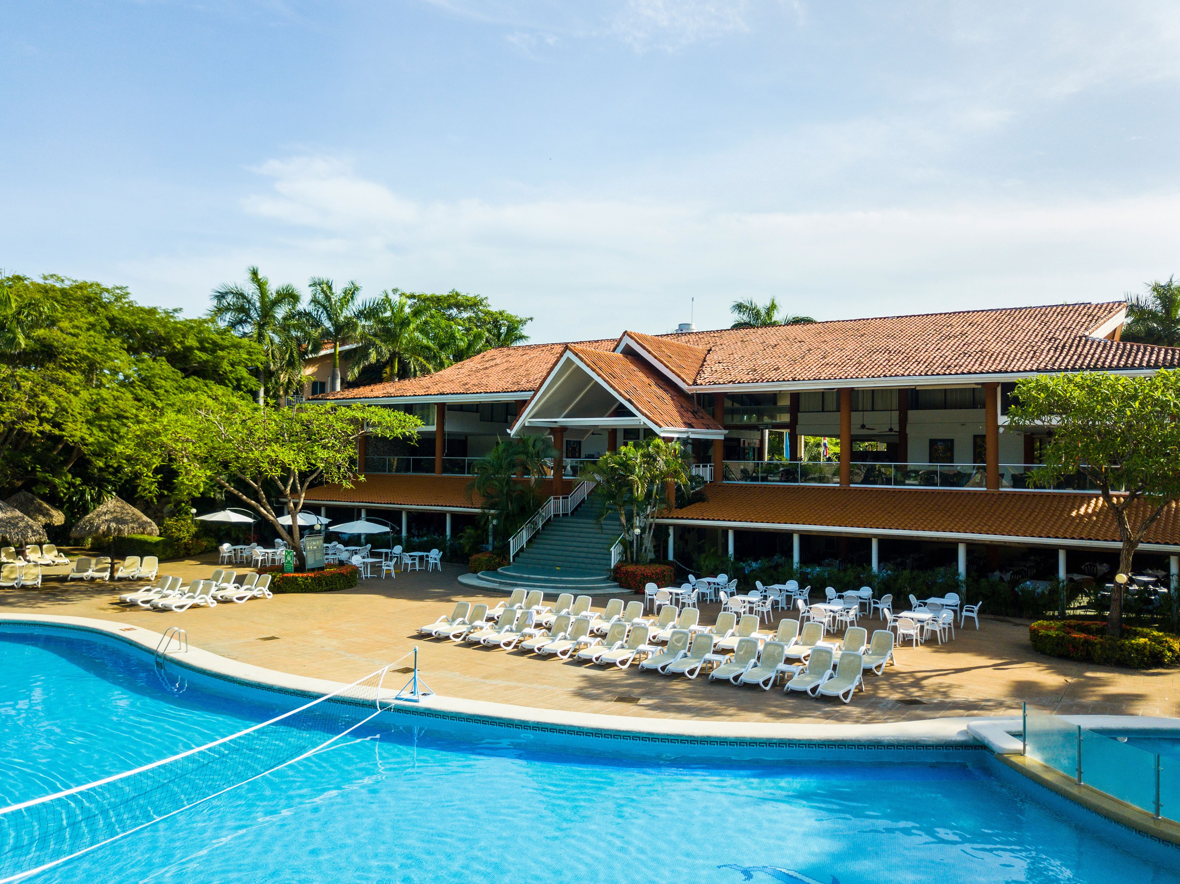 A resort with a swimming pool, lounge chairs, and a two-story building with a tiled roof.