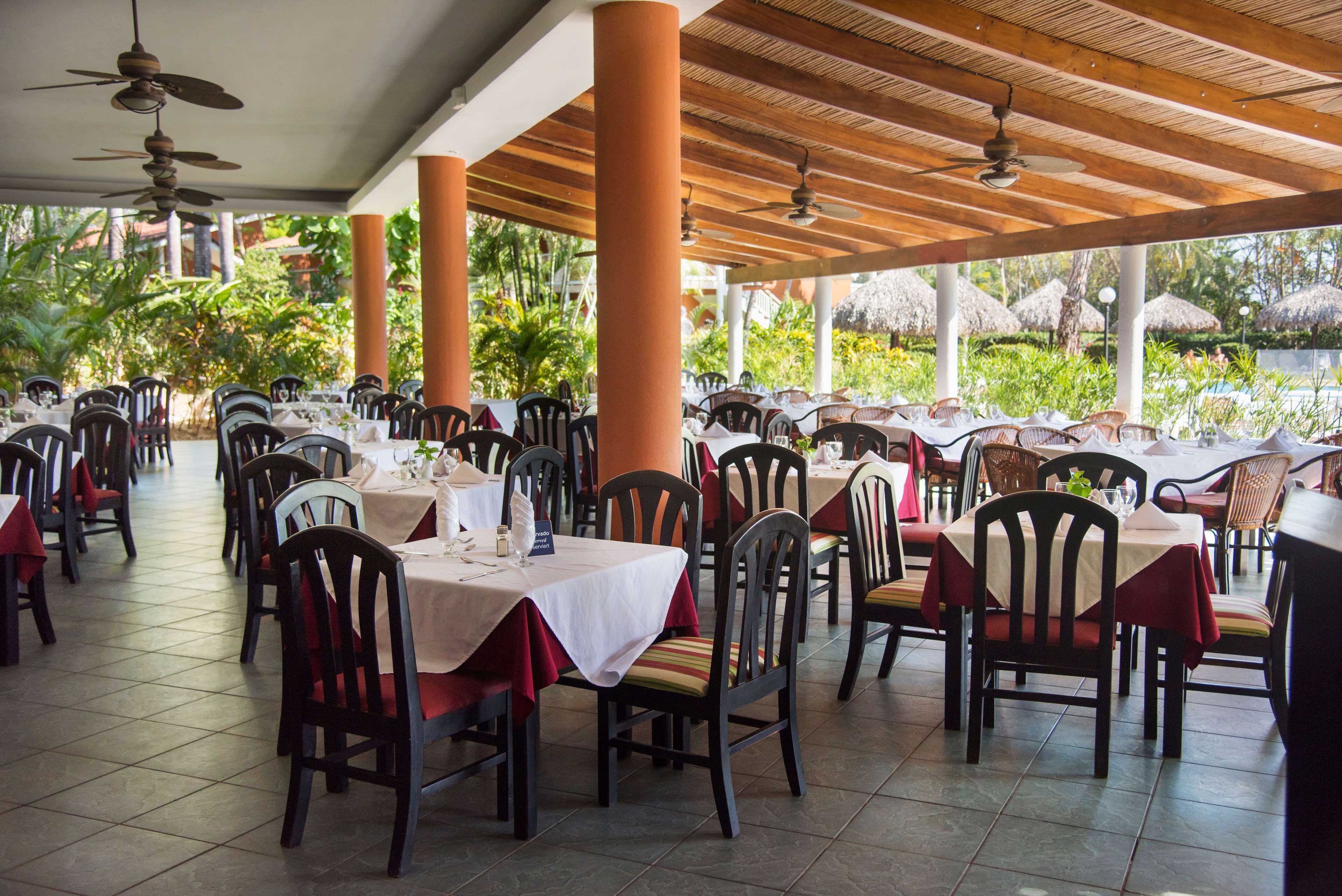 A covered outdoor dining area with multiple tables and chairs, surrounded by lush greenery and thatched umbrellas.