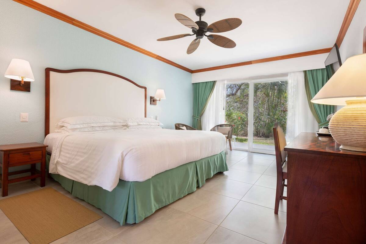 Un dormitorio con cama, mesitas de noche, ventilador de techo, ventana con cortinas y un comedor con mesa y sillas.
