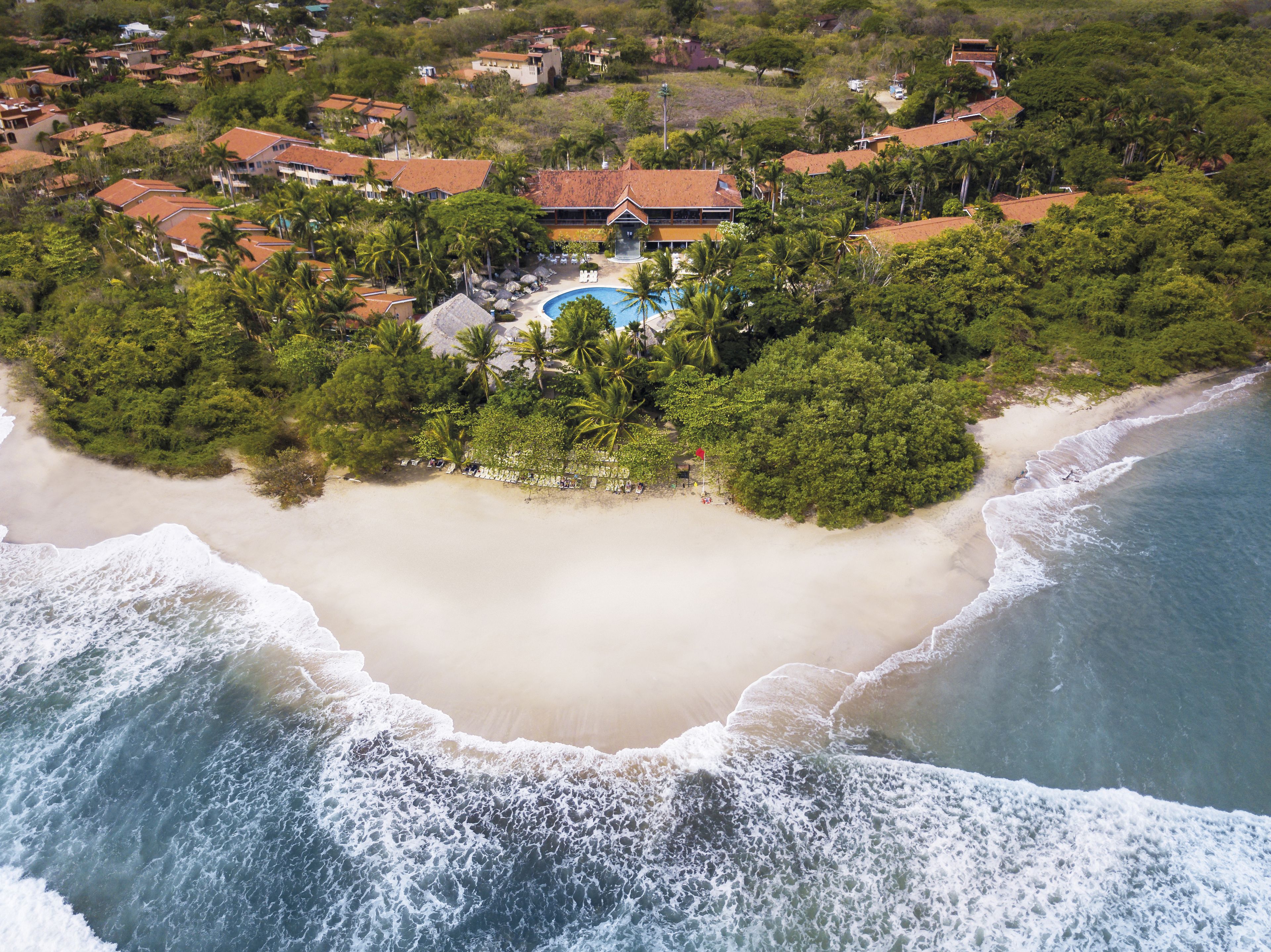 An aerial view of a coastal resort with a large sandy beach, a swimming pool, and lush greenery.
