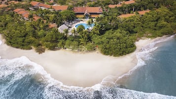 An aerial view of a coastal resort with a large sandy beach, a swimming pool, and lush greenery.