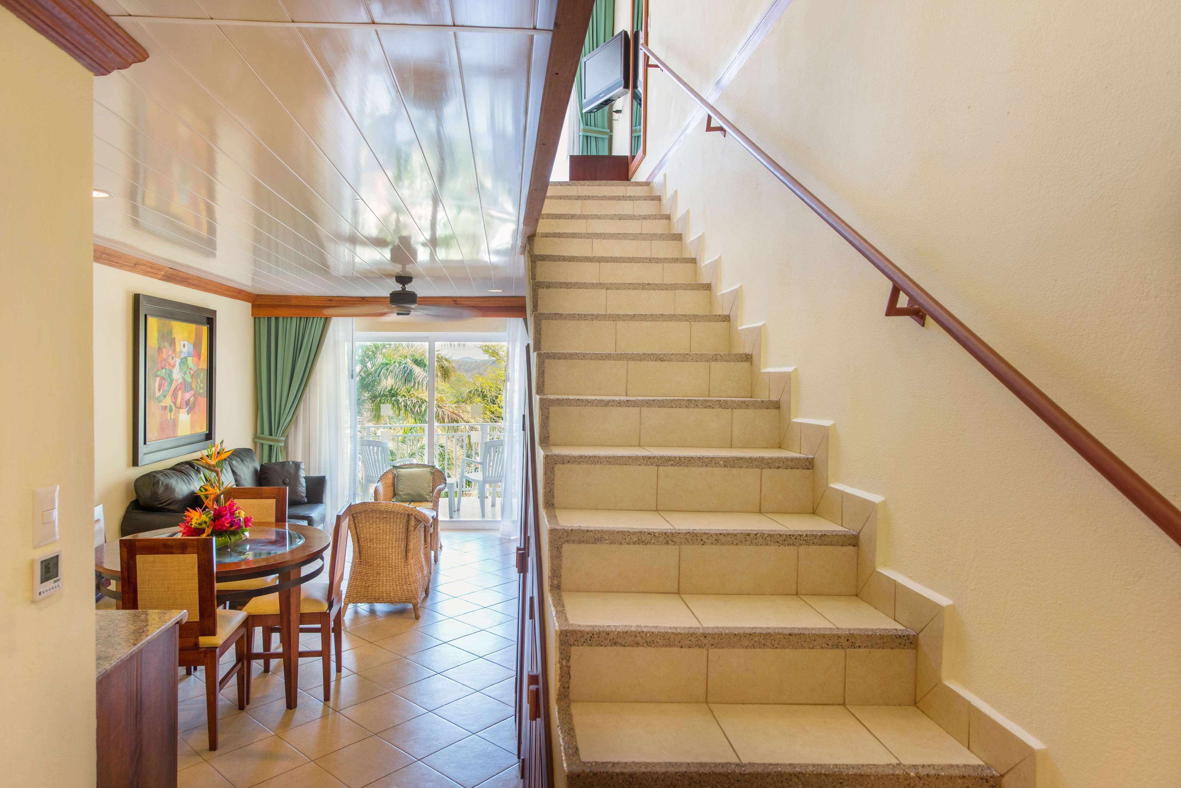 Una escalera con barandilla, un comedor con mesa y sillas, y vista al exterior por una puerta.