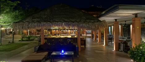 A thatched-roof outdoor bar area with seating and a pool table, surrounded by wooden decking and potted plants.