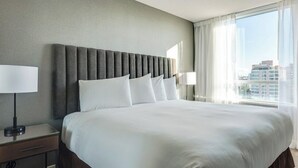 A hotel room with a large bed, a bedside table with a lamp, and a view of the city through large windows.