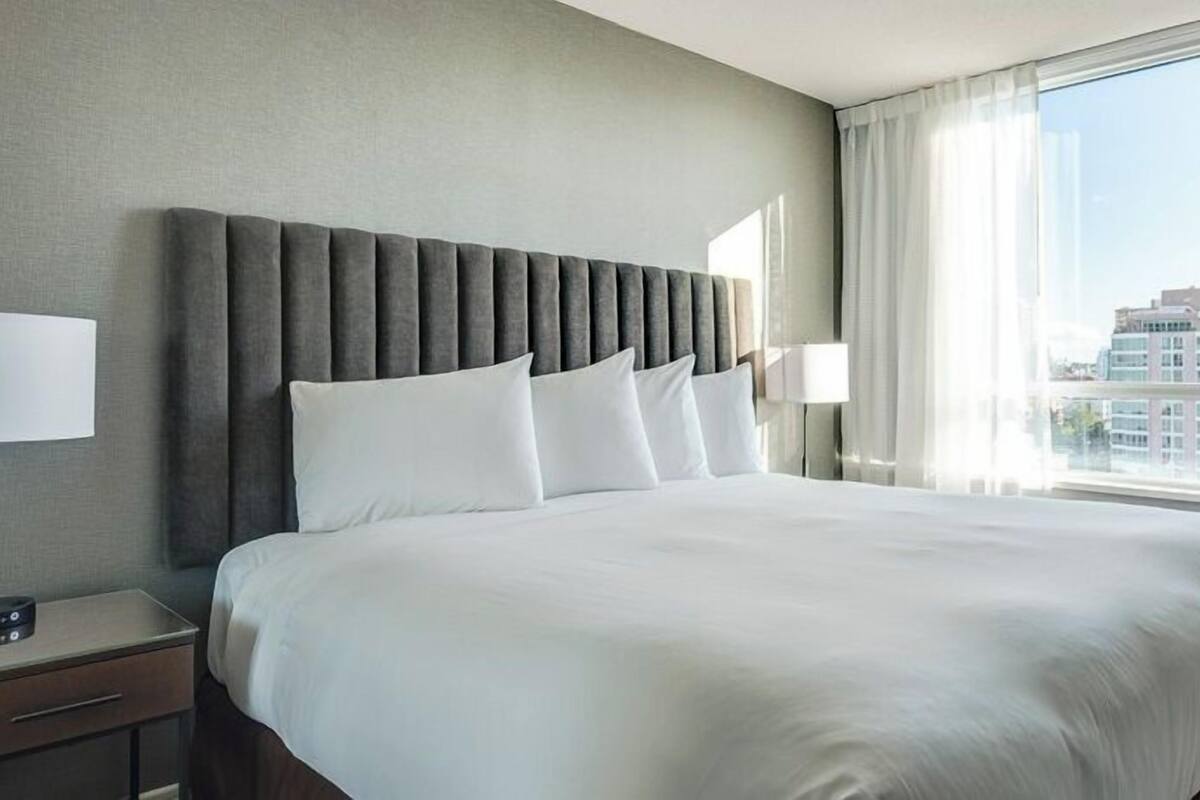 A hotel room with a large bed, a bedside table with a lamp, and a view of the city through large windows.