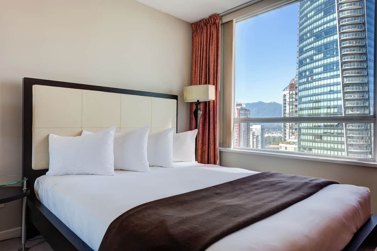 A hotel room with a large bed, a nightstand, a lamp, and a window with a city view.