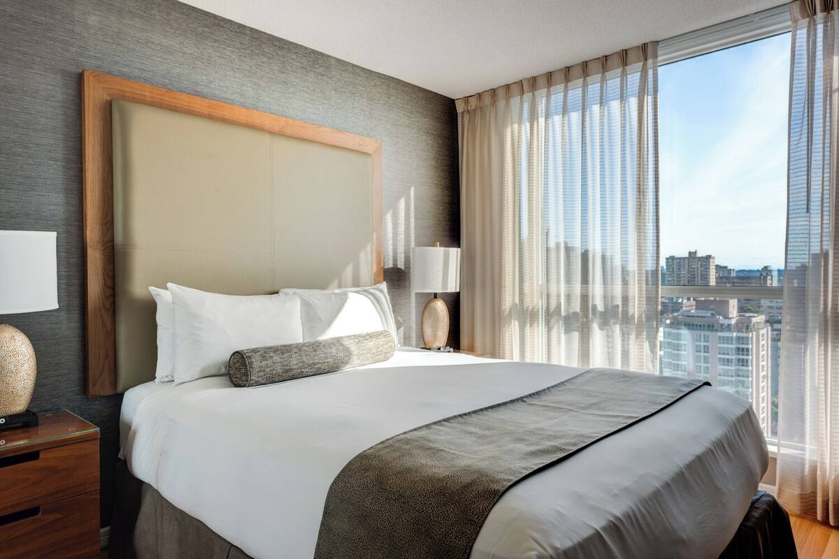A hotel room with a large bed, a nightstand, a lamp, and a view of the city through sheer curtains.