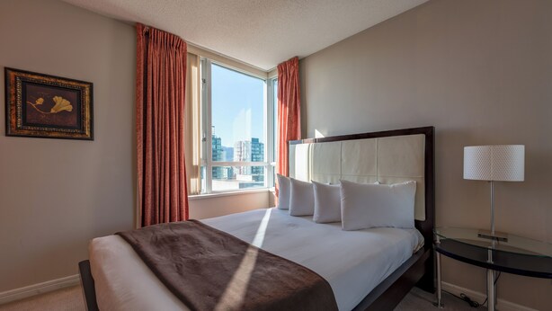 A hotel room with a large bed, a bedside table with a lamp, a window with curtains, and a framed picture on the wall. - Carmana Hotel & Suites (Vancouver)