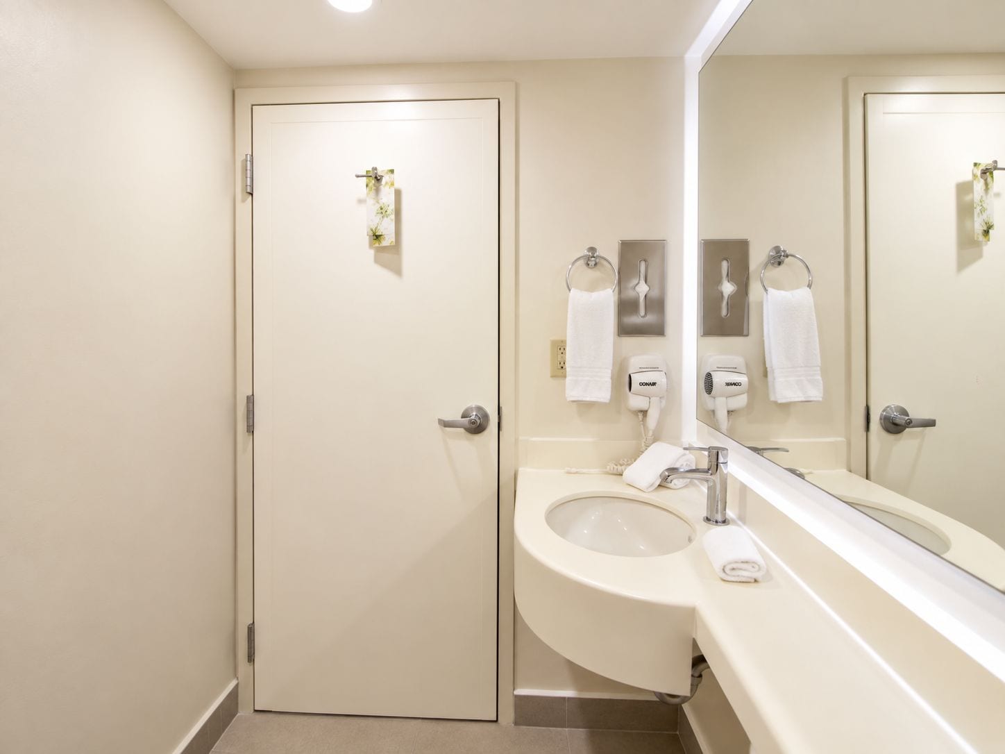 A bathroom with a toilet, sink, and a large mirror. There are two towel racks and two hair dryers on the wall.
