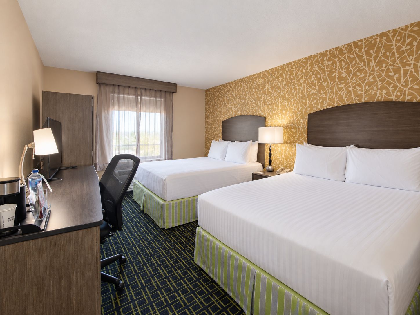 Quarto de hotel com duas camas, uma escrivaninha com cadeira, televisão e janela com cortinas.