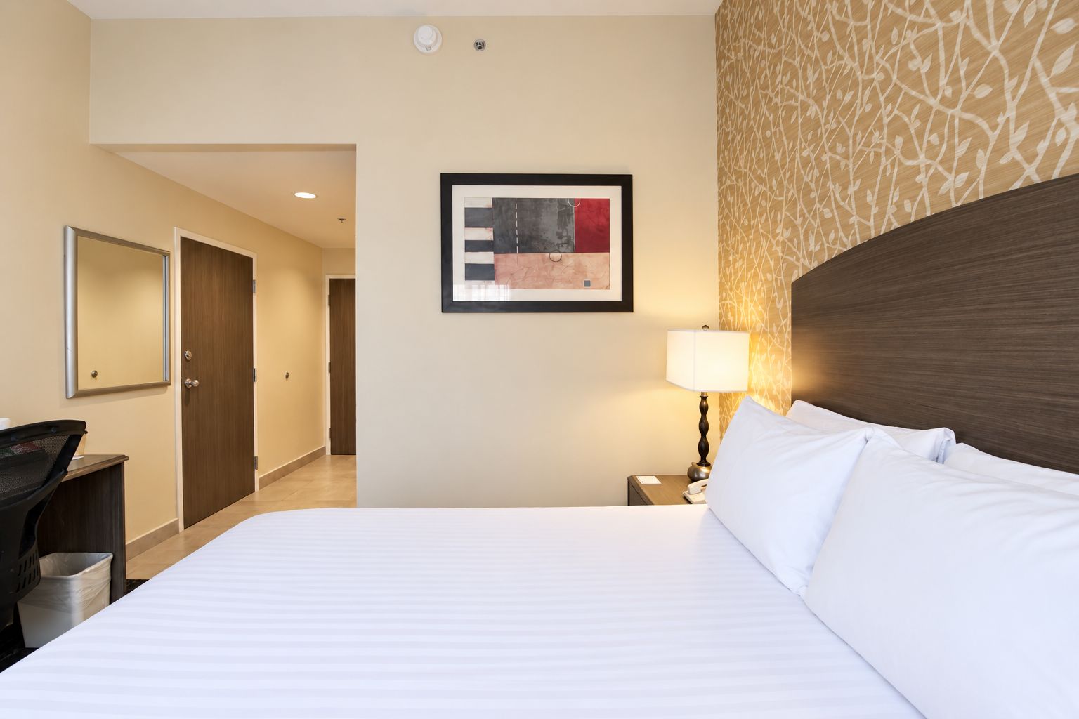 A hotel room with a bed, a desk, a lamp, a framed artwork, and a patterned wall.