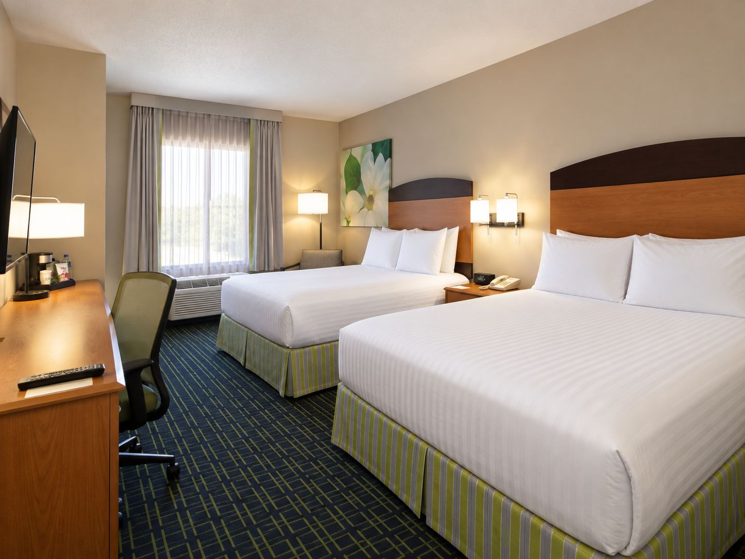 A hotel room with two beds, a desk, a chair, a lamp, a telephone, and a painting on the wall.
