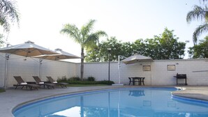 Ein Swimmingpool mit Liegestühlen und Sonnenschirmen, umgeben von einer weißen Kachelwand und Grünpflanzen.