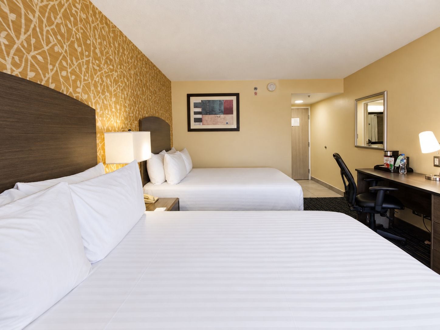 A hotel room with two beds, a desk, a chair, a lamp, a clock, and a framed picture on the wall.