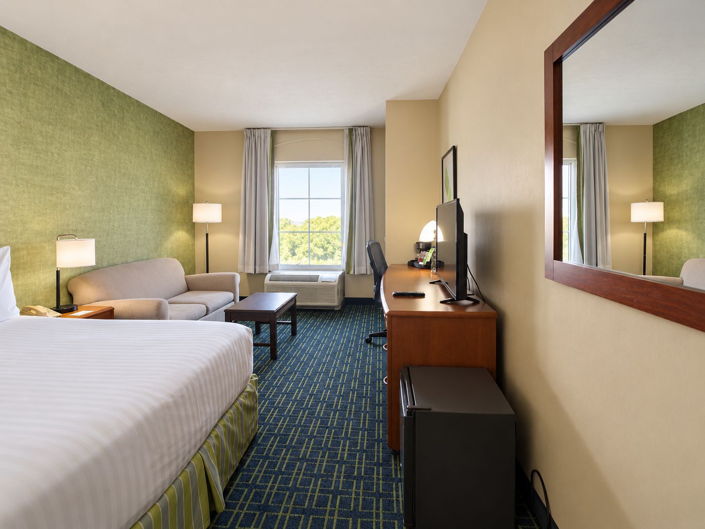 A hotel room with a bed, a desk, a chair, a small refrigerator, and a window with curtains.