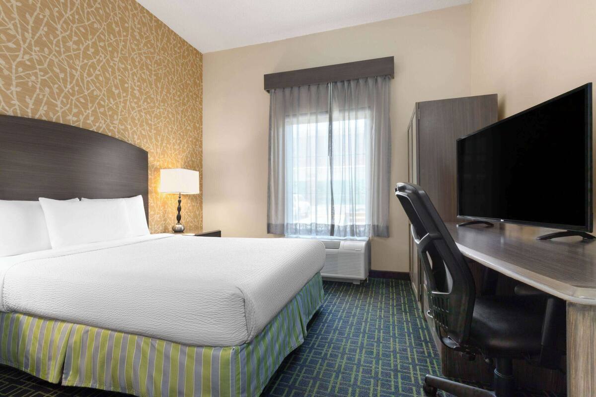 Habitación de hotel con una cama grande, un escritorio, un televisor de pantalla plana, una lámpara y una ventana con cortinas.