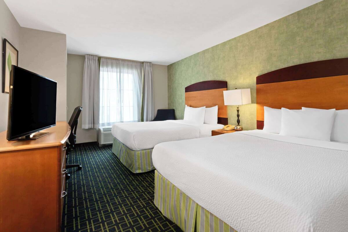 Habitación de hotel con dos camas, un escritorio, un televisor de pantalla plana, una silla, una lámpara y una ventana con cortinas.