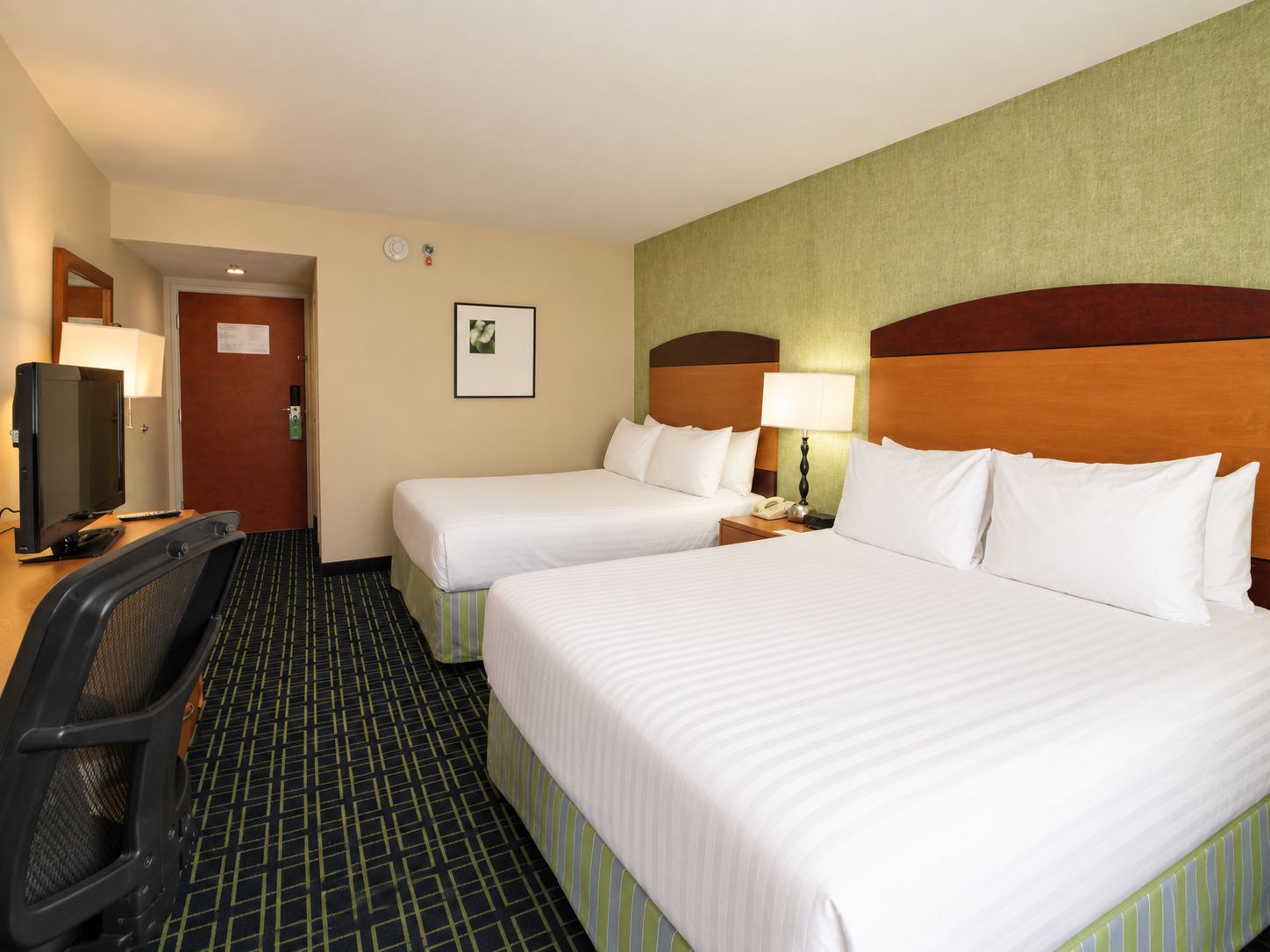 A hotel room with two beds, a desk with a computer, a chair, a television, and a wall-mounted lamp.