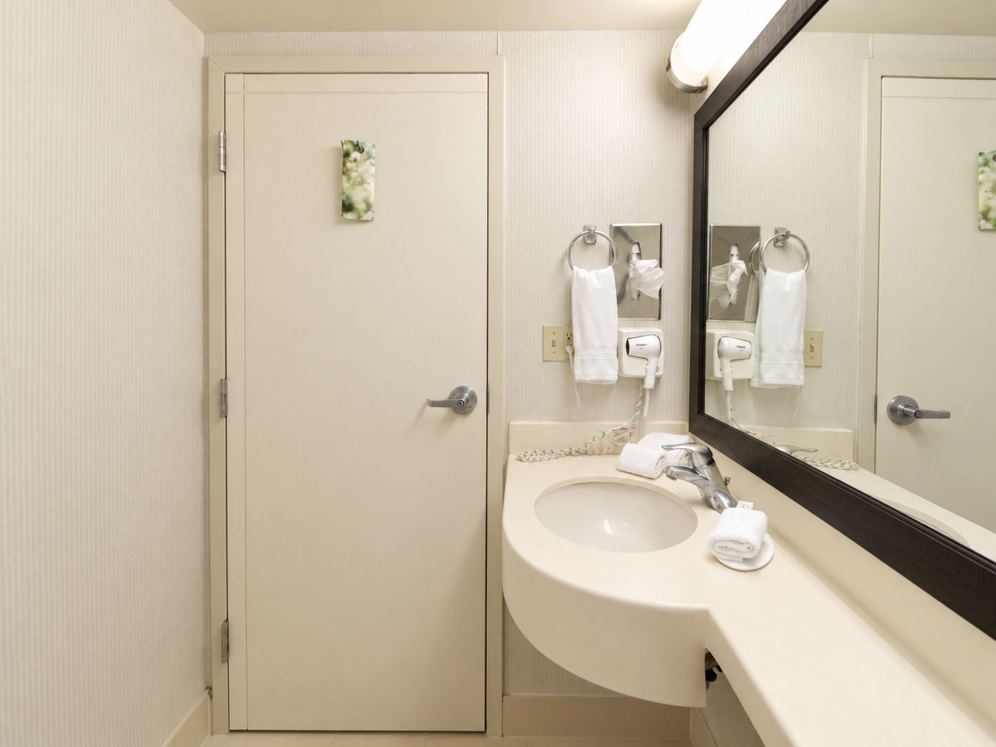 A hotel bathroom with a white sink, a mirror, and a door with a small floral decoration.