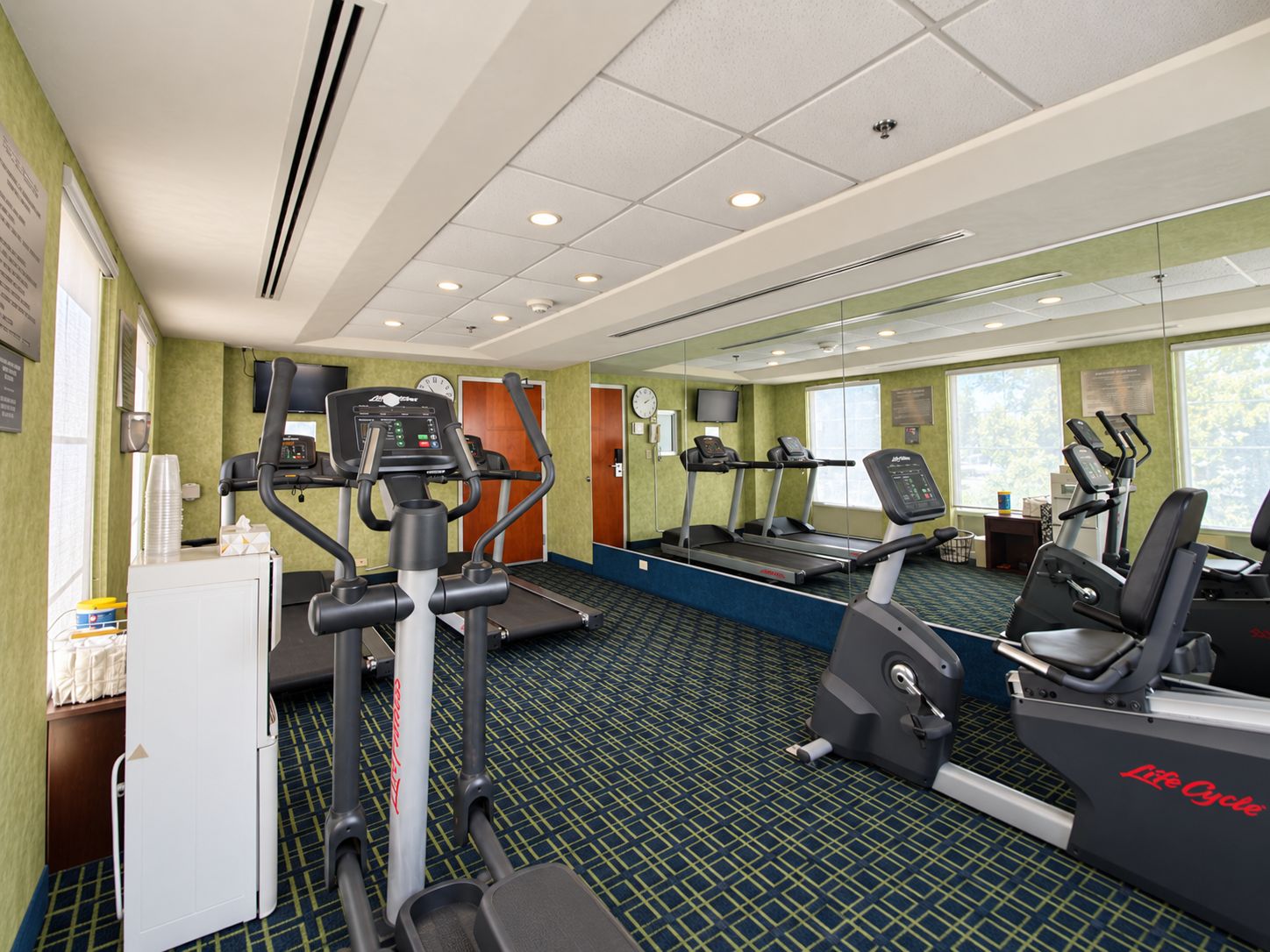 A hotel gym with cardio machines, a mirrored wall, and a green carpeted floor.