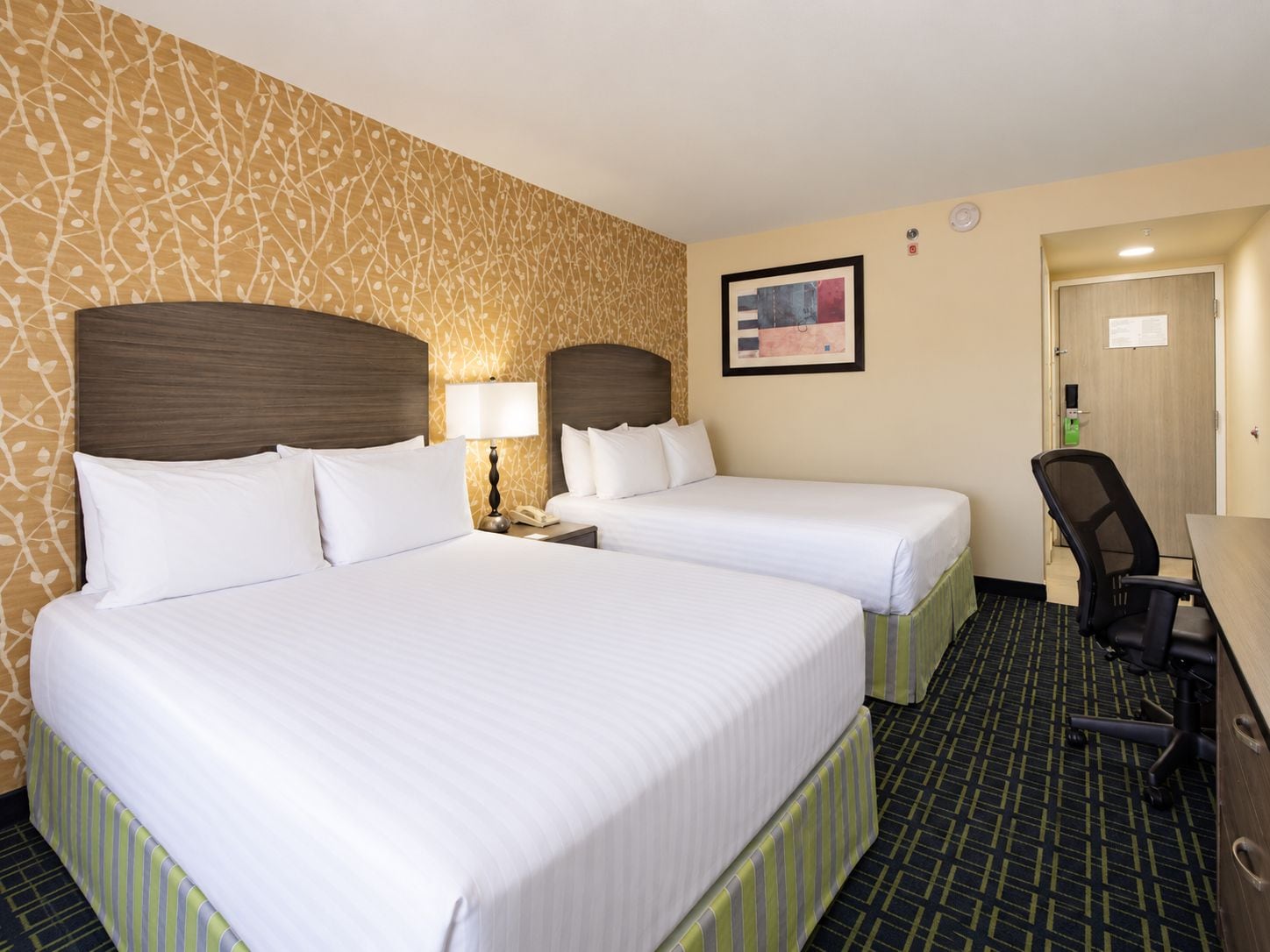 A hotel room with two beds, a desk, a chair, and a patterned wall.